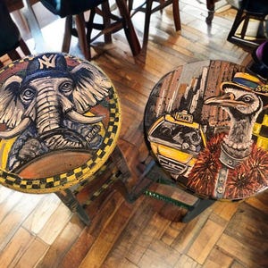 Puede incluir: Dos taburetes redondos con retratos de animales pintados. Uno presenta un elefante con gorra de béisbol, el otro un avestruz en un taxi. Ambos tienen un borde a cuadros y están sobre un suelo de madera.