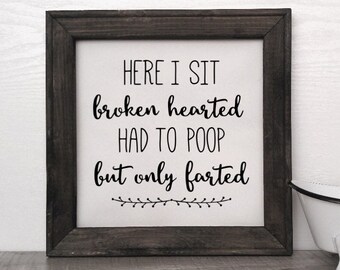 Peut inclure: Une pancarte en bois rustique encadrée avec le texte "HERE I SIT broken hearted HAD TO POOP but only farted" en écriture noire. Une petite baignoire blanche avec des garnitures noires est posée sur une surface en bois.