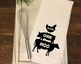 May include: White kitchen towel featuring black silhouettes of a cow, pig, and chicken, with the words "MOO", "OINK", and "CLUCK". A silver whisk is placed beside the towel. The towel is on a white wooden tray.