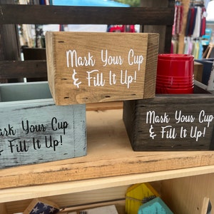 May include: Three wooden boxes with the text "Mark Your Cup & Fill It Up!" painted on them. The boxes are painted in different colors: blue, brown, and natural wood. The boxes are displayed on a wooden shelf.