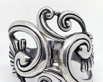 May include: A silver bracelet with a delicate, intricate design of swirling, openwork patterns. The bracelet is made of polished metal and has a hinged closure.