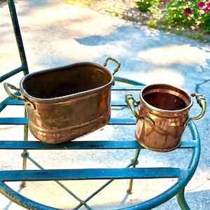 May include: Two copper vessels with brass handles. One is an oval-shaped container with two side handles, and the other is a smaller, cylindrical pot with two ornate brass handles on opposite sides. Both items feature a polished metallic finish.