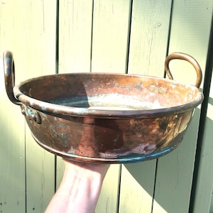May include: A vintage copper bowl with two curved handles. The bowl has a wide, shallow design and a rich, aged patina with areas of verdigris. The bowl is held up against a light green wooden background.