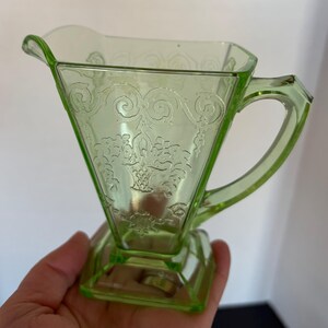 May include: A vintage, light green glass creamer with a decorative floral pattern. The creamer has a triangular body, a handle, and a square base. The glass is translucent, and the design is etched into the surface.