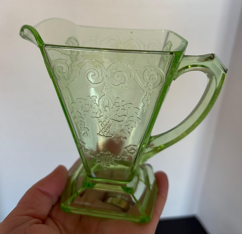 May include: A vintage, light green glass creamer with a decorative floral pattern. The creamer has a triangular body, a handle, and a square base. The glass is translucent, and the design is etched into the surface.
