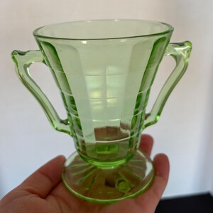 May include: A light green glass sugar bowl with two handles. The bowl has a square pattern design and sits on a round base. The handles are curved and the glass has a slight glow.