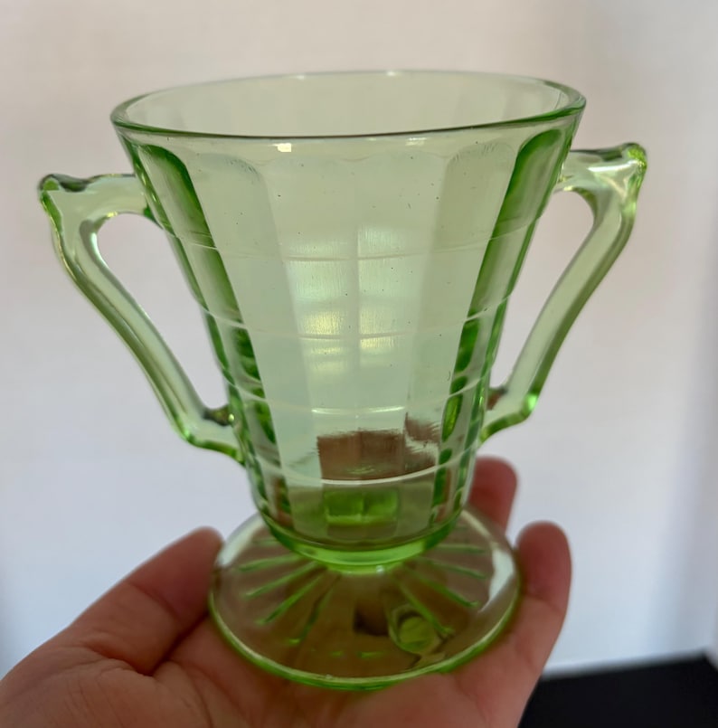 May include: A light green glass sugar bowl with two handles. The bowl has a square pattern design and sits on a round base. The handles are curved and the glass has a slight glow.