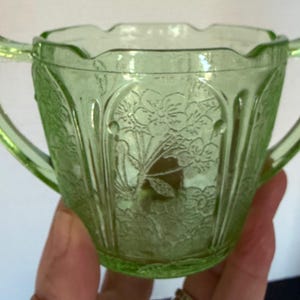 May include: A vintage green glass sugar bowl with two handles. The bowl features a floral pattern and a scalloped rim. The glass has a slight glow, suggesting it may be uranium glass. A decorative piece for serving sugar.
