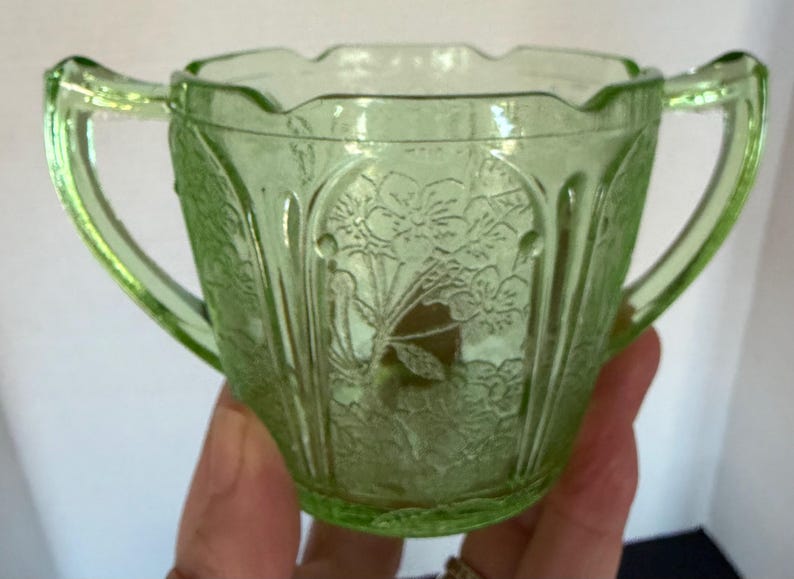 May include: A vintage green glass sugar bowl with two handles. The bowl features a floral pattern and a scalloped rim. The glass has a slight glow, suggesting it may be uranium glass. A decorative piece for serving sugar.