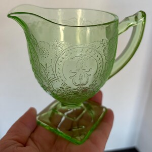 May include: A light green glass creamer with a decorative floral and medallion pattern. The creamer has a handle and a spout for pouring. The base is square and the creamer is held in a hand.
