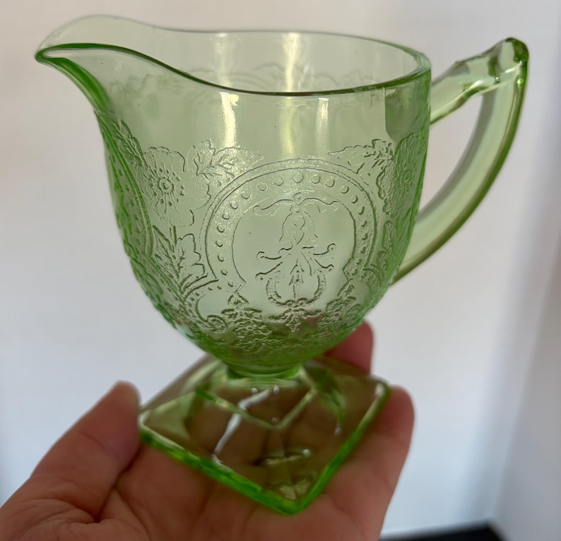 May include: A light green glass creamer with a decorative floral and medallion pattern. The creamer has a handle and a spout for pouring. The base is square and the creamer is held in a hand.
