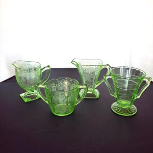 May include: Four pieces of green Depression glass, including a creamer, sugar bowl, and two different styles of handled cups. The glass has etched patterns and a vibrant, translucent appearance. The set is displayed on a dark surface.