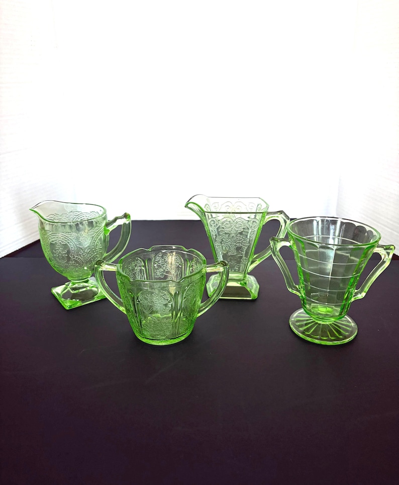 May include: Four pieces of green Depression glass, including a creamer, sugar bowl, and two different styles of handled cups. The glass has etched patterns and a vibrant, translucent appearance. The set is displayed on a dark surface.