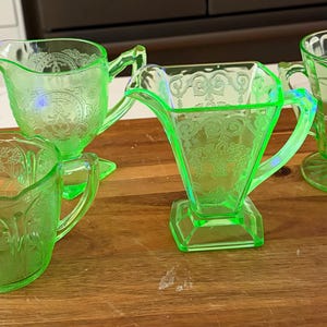 May include: Four pieces of vintage green depression glass, including a creamer, sugar bowl, and two different shaped handled glasses. The glassware is displayed on a wooden surface. The pieces have etched floral and geometric patterns.