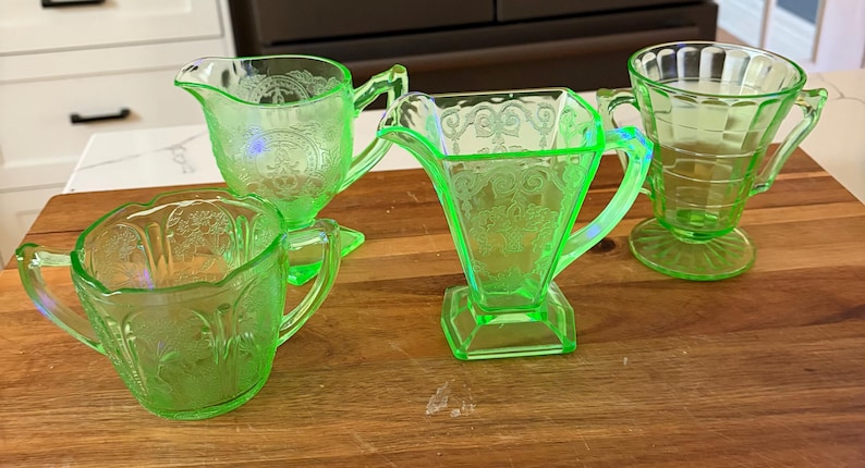 May include: Four pieces of vintage green depression glass, including a creamer, sugar bowl, and two different shaped handled glasses. The glassware is displayed on a wooden surface. The pieces have etched floral and geometric patterns.