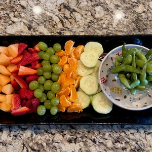 Könnte beinhalten: Eine schwarze rechteckige Platte mit einer Auswahl an Früchten, die in Reihen angeordnet sind. Die Früchte umfassen Cantaloupe, Erdbeeren, grüne Trauben, Mandarinen und Gurken. Eine kleine weiße Schüssel mit grünen Edamame-Bohnen befindet sich auf der Platte.