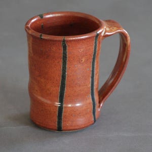 Könnte beinhalten: Eine rote Keramiktasse mit schwarzen Streifen. Die Tasse hat einen Henkel und ist im rustikalen Stil gehalten.