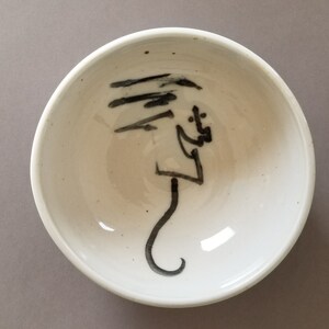 Puede incluir: Un cuenco de cerámica blanca con un diseño abstracto negro pintado en el interior. El diseño se asemeja a un árbol estilizado con un tronco largo y curvo.