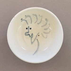 May include: A white ceramic bowl with a blue and white hand-painted design of a face with a whimsical hairstyle.