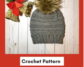 May include: Gray crochet beanie with a brown faux fur pom pom. The beanie is on a white wooden background with gold bells and red ribbon.