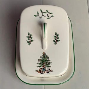 May include: White ceramic Christmas themed butter dish with a green trim. The lid features a green Christmas tree with ornaments and a sprig of holly. The base has a green trim and a sprig of holly.