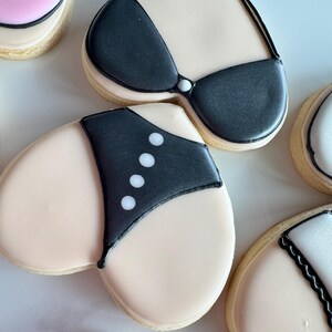 May include: Heart-shaped sugar cookies decorated with black and white icing, depicting a swimsuit and bra. The cookies are on a white surface.