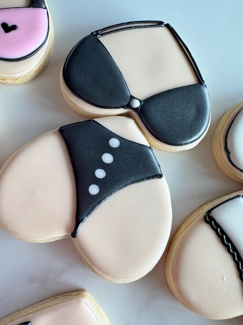 May include: Heart-shaped sugar cookies decorated with black and white icing, depicting a swimsuit and bra. The cookies are on a white surface.