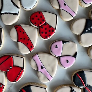 May include: Assortment of heart-shaped cookies decorated with various lingerie designs. The cookies are decorated with red, pink, black, and white icing. Some cookies have polka dots.
