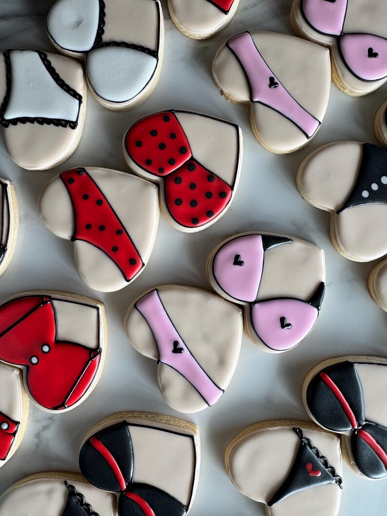 May include: Assortment of heart-shaped cookies decorated with various lingerie designs. The cookies are decorated with red, pink, black, and white icing. Some cookies have polka dots.