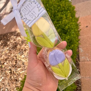 May include: A clear plastic bag with a white ribbon and a card that says "Happy Mother's Day". Inside the bag are two sugar cookies decorated with yellow and purple icing to look like tulips.