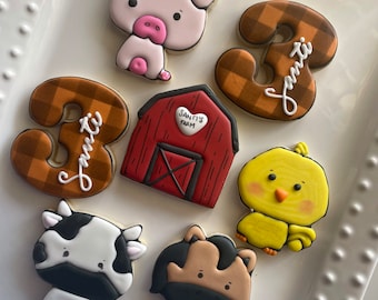May include: A set of seven decorated sugar cookies featuring a pig, a cow, a horse, a chick, a red barn, and two brown and white checkered cookies with the number "3" and the name "Santi" written on them.