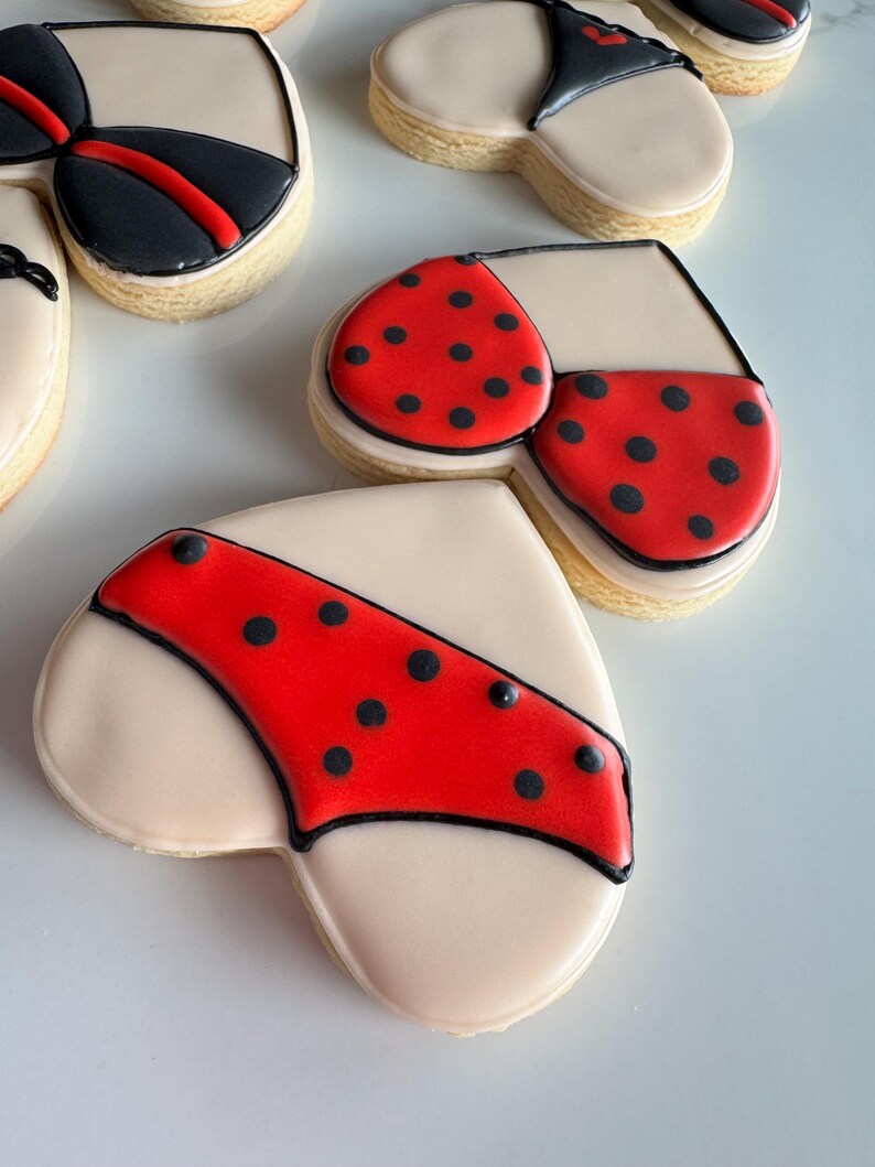 May include: Heart-shaped cookies decorated with red and black icing, resembling lingerie. The cookies have a white background and black outlines.
