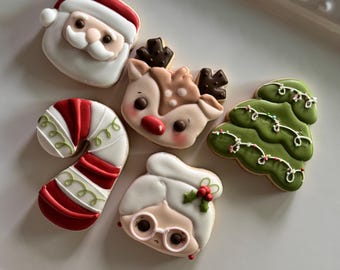 May include: A collection of Christmas-themed cookies, including Santa Claus, a reindeer, a Christmas tree, Mrs. Claus, and a candy cane. The cookies are decorated with white, red, green, and brown icing, and are arranged on a white platter.