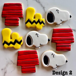 May include: A set of nine decorated sugar cookies featuring the characters Snoopy and Woodstock from the Peanuts comic strip. The cookies are decorated with red, yellow, black, and white icing. The cookies are arranged in a pattern on a white surface. Design 2.