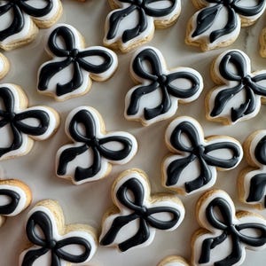 May include: A collection of sugar cookies decorated with black icing in the shape of bows. The cookies are arranged on a white surface.