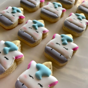 May include: A collection of decorated sugar cookies shaped like unicorn heads. The cookies are decorated with white, blue, pink, and gold icing.