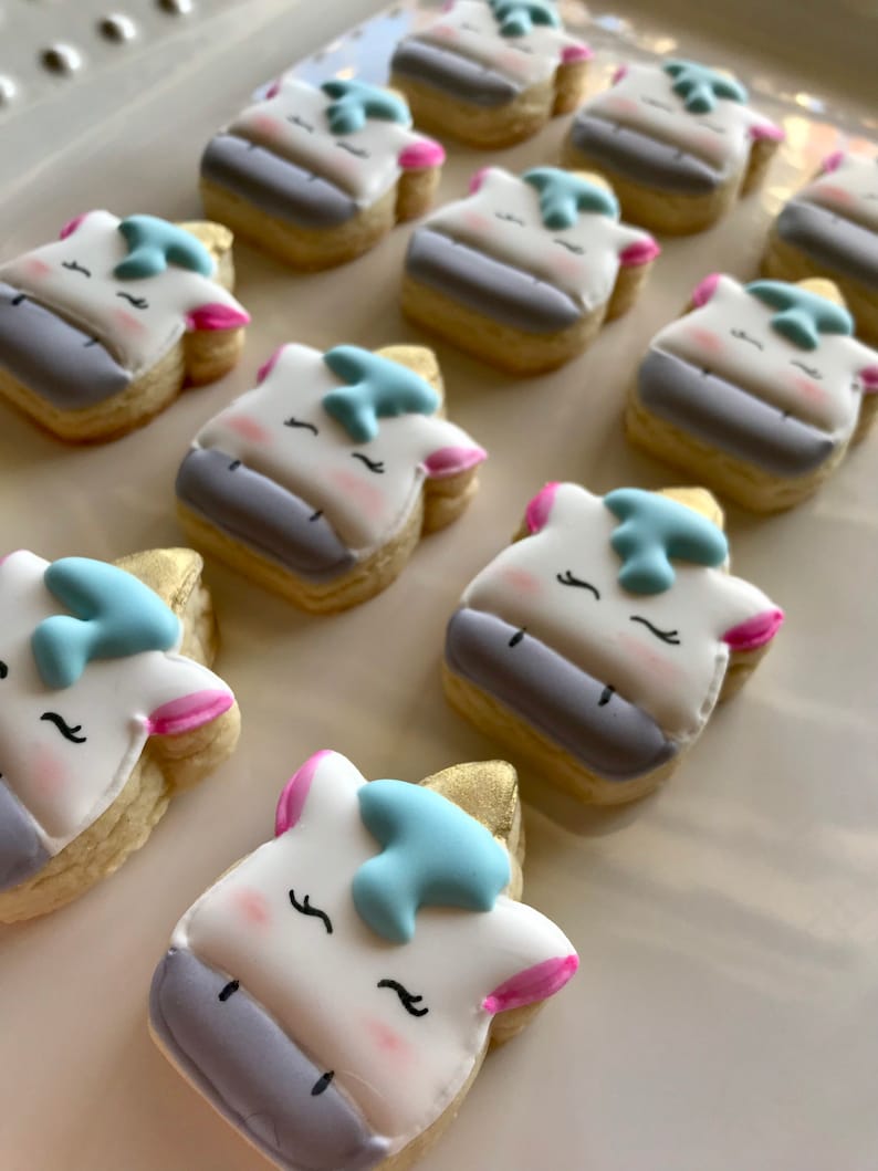 May include: A collection of decorated sugar cookies shaped like unicorn heads. The cookies are decorated with white, blue, pink, and gold icing.