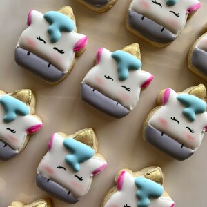 May include: A collection of nine unicorn-shaped sugar cookies decorated with white, pink, blue, and gold icing. The cookies have a white base with a grey mane and tail, and each cookie has a gold horn and blue eyes.