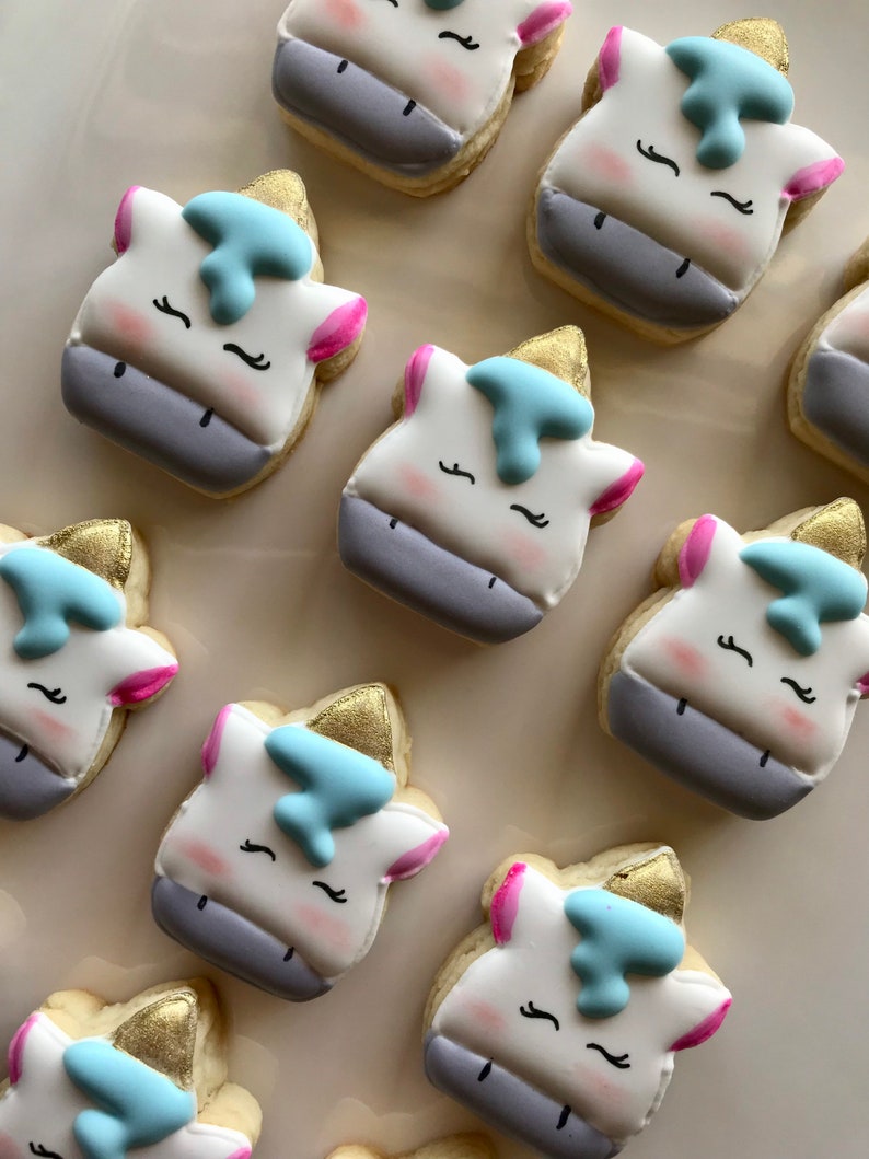 May include: A collection of nine unicorn-shaped sugar cookies decorated with white, pink, blue, and gold icing. The cookies have a white base with a grey mane and tail, and each cookie has a gold horn and blue eyes.