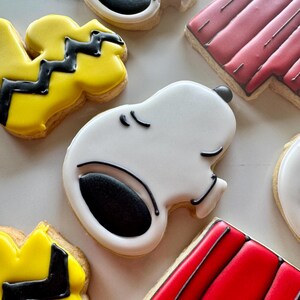 May include: A set of six decorated sugar cookies featuring characters from the Peanuts comic strip. The cookies are decorated with white, black, yellow, and red icing. The characters include Snoopy, Woodstock, and Charlie Brown.