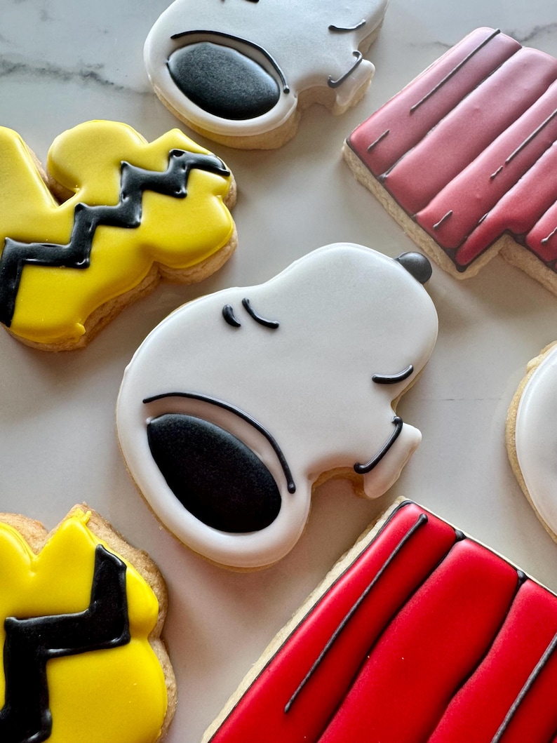 May include: A set of six decorated sugar cookies featuring characters from the Peanuts comic strip. The cookies are decorated with white, black, yellow, and red icing. The characters include Snoopy, Woodstock, and Charlie Brown.