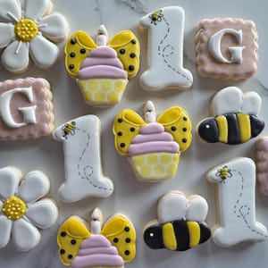 May include: Decorated sugar cookies in a variety of shapes, including daisies, cupcakes, bees, the number one, and squares with the letter "G". The cookies are decorated with white, yellow, black, and pink icing.
