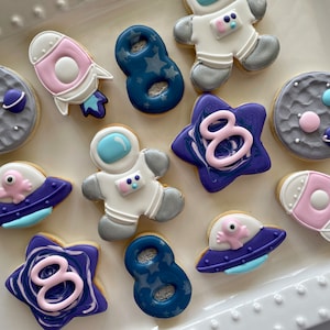 May include: A set of ten decorated sugar cookies with an outer space theme. The cookies are decorated with purple, blue, pink, and gray icing. The cookies feature images of planets, stars, spaceships, and astronauts.