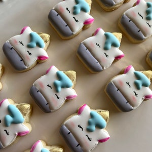 May include: A collection of nine unicorn-themed sugar cookies decorated with white, pink, blue, and gold icing. The cookies are arranged in a grid pattern on a white surface.