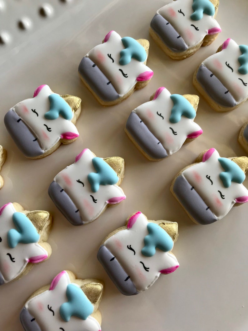 May include: A collection of nine unicorn-themed sugar cookies decorated with white, pink, blue, and gold icing. The cookies are arranged in a grid pattern on a white surface.