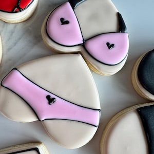 May include: Heart-shaped sugar cookies decorated with pink and black icing to resemble lingerie. The cookies are on a white marble surface.
