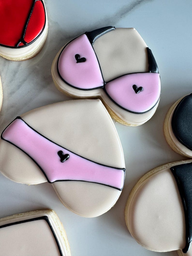 May include: Heart-shaped sugar cookies decorated with pink and black icing to resemble lingerie. The cookies are on a white marble surface.