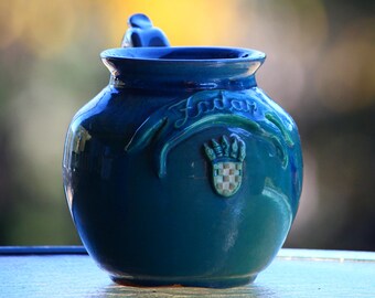 Puede incluir: Una jarra de cerámica azul verdosa con asa y un diseño decorativo con un escudo y la palabra "Fador".