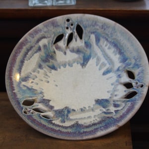 Puede incluir: Un plato de cerámica con un diseño único. El plato presenta un centro blanco y un borde iridiscente azul y morado. Tiene formas recortadas decorativas alrededor del borde, lo que aumenta su atractivo artístico. El plato se exhibe sobre una superficie de madera.
