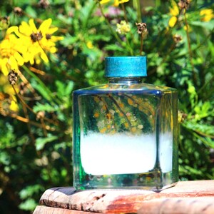 Puede incluir: Una botella de vidrio transparente y cuadrada con una tapa turquesa, llena de un líquido blanco. La botella está sobre una superficie de madera, con flores amarillas y follaje verde de fondo. El contenido de la botella parece opaco.